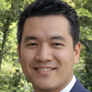 Simon smiling outdoors, dressed in a suit, with short black hair.